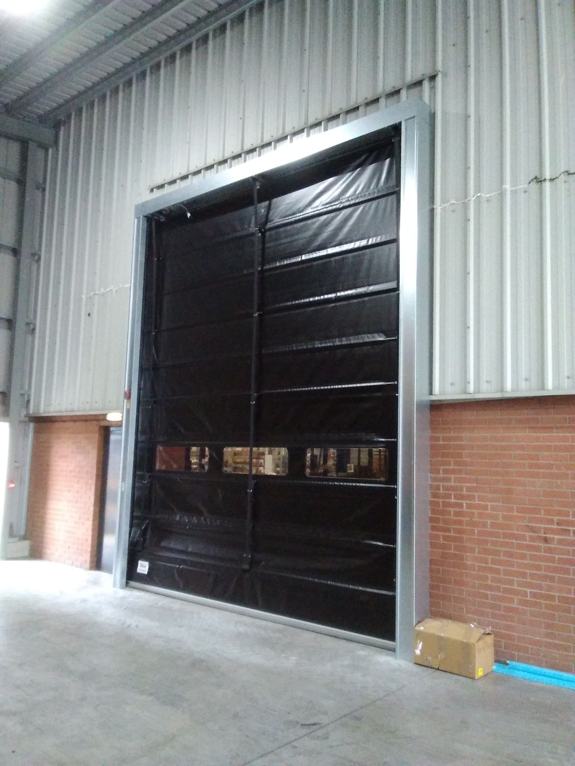 A large industrial black roller shutter is installed in a warehouse wall, with a small cardboard box on the floor nearby.