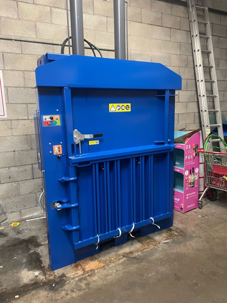 A large blue industrial baler stands against a breeze block wall, with warning labels and control buttons visible on its side. Nearby are a ladder, a trolley, and a pink vending machine.