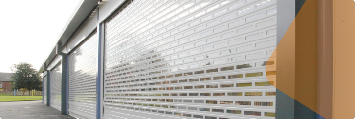 Row of partially open, white perforated roller shutter doors on a modern commercial or industrial building, with a grassy area and trees visible in the background.