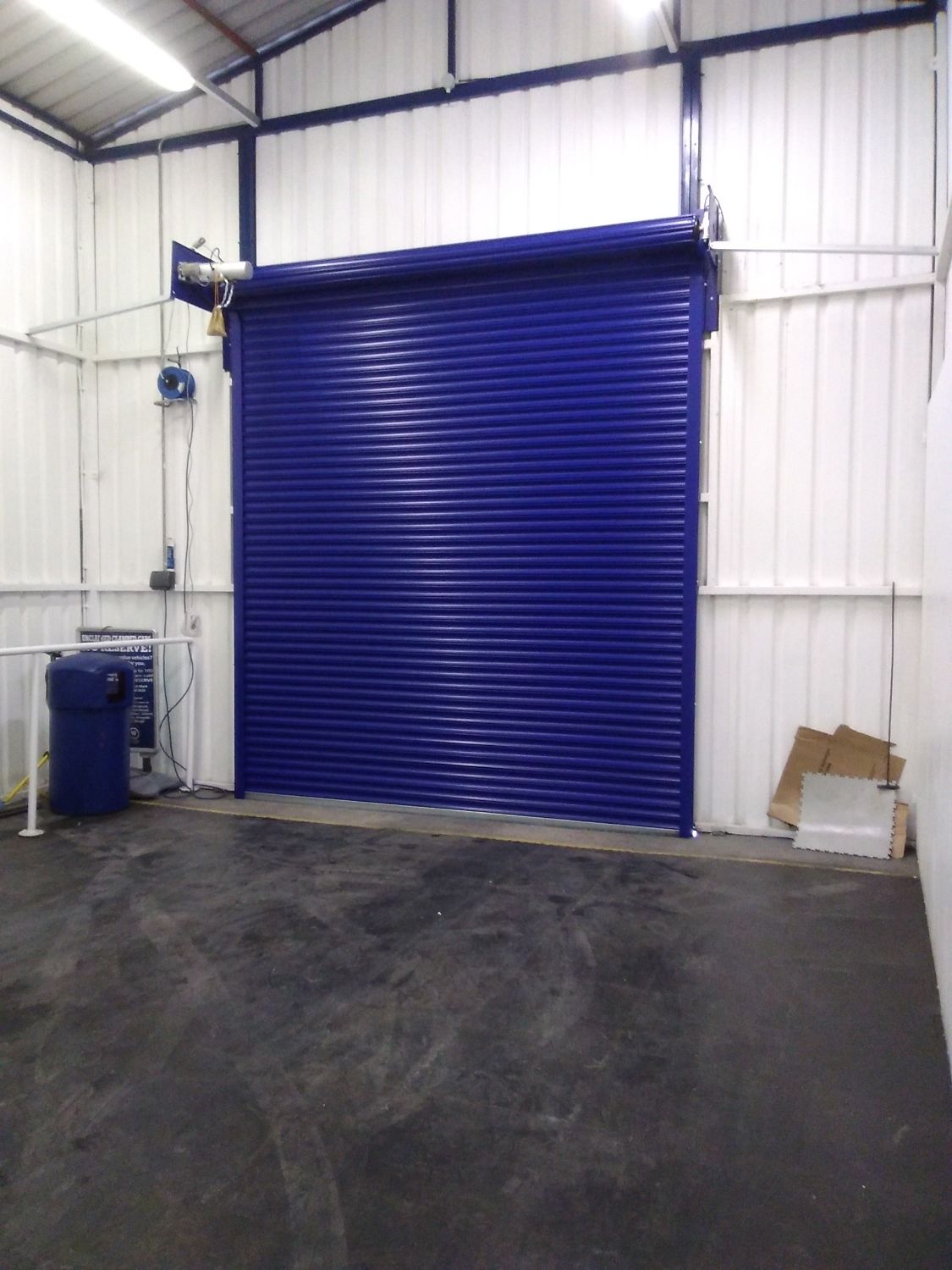 Blue metal roller shutter door in a clean, empty industrial warehouse with white corrugated walls and concrete floor; equipment and cardboard are placed in the corners.