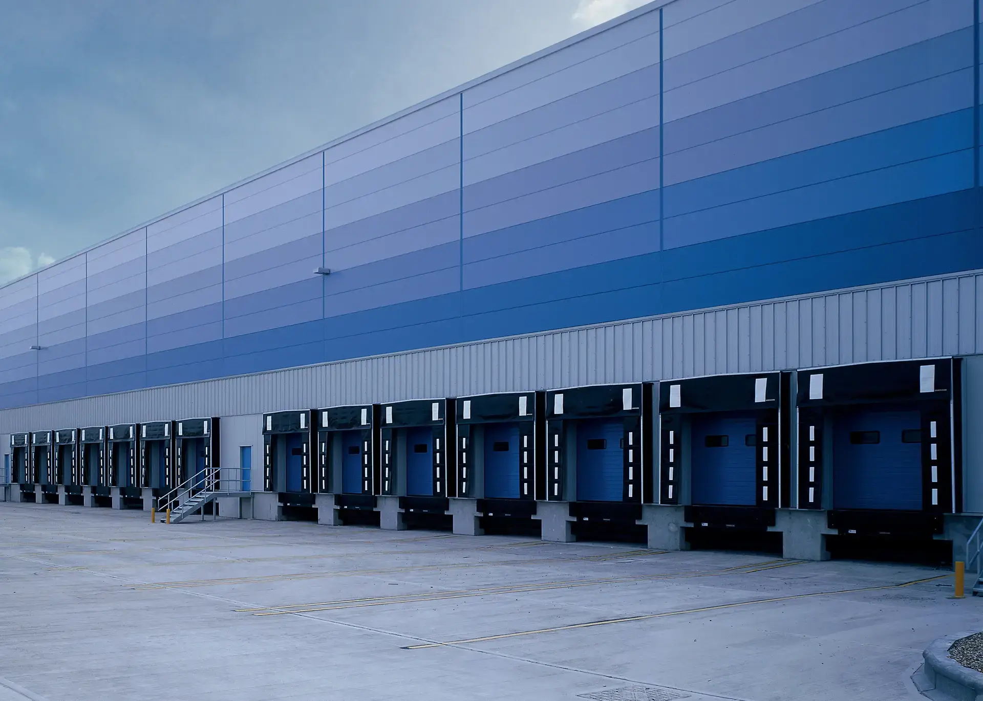 A row of closed loading bay doors on the side of a blue industrial warehouse building under a cloudy sky.
