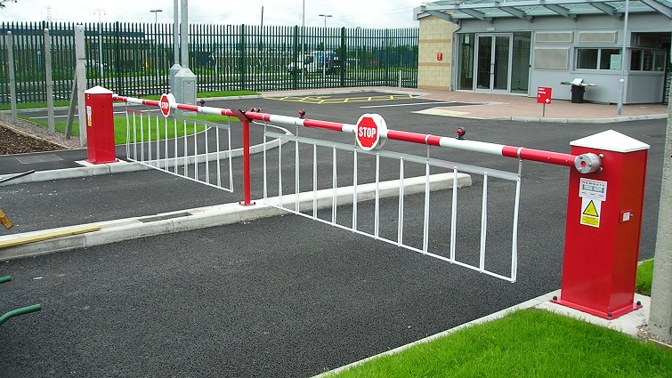 A red and white security barrier gate with "STOP" signs blocks vehicle access to a paved area near a building and a fenced perimeter.