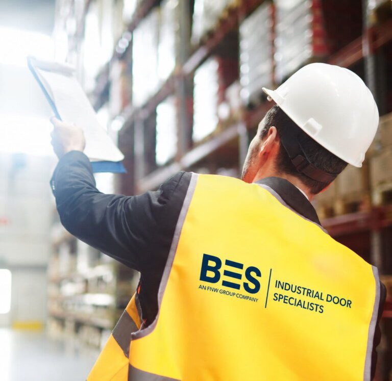 Two workers in safety vests and helmets inspect shelves in a warehouse, with one holding a clipboard and pointing upwards.