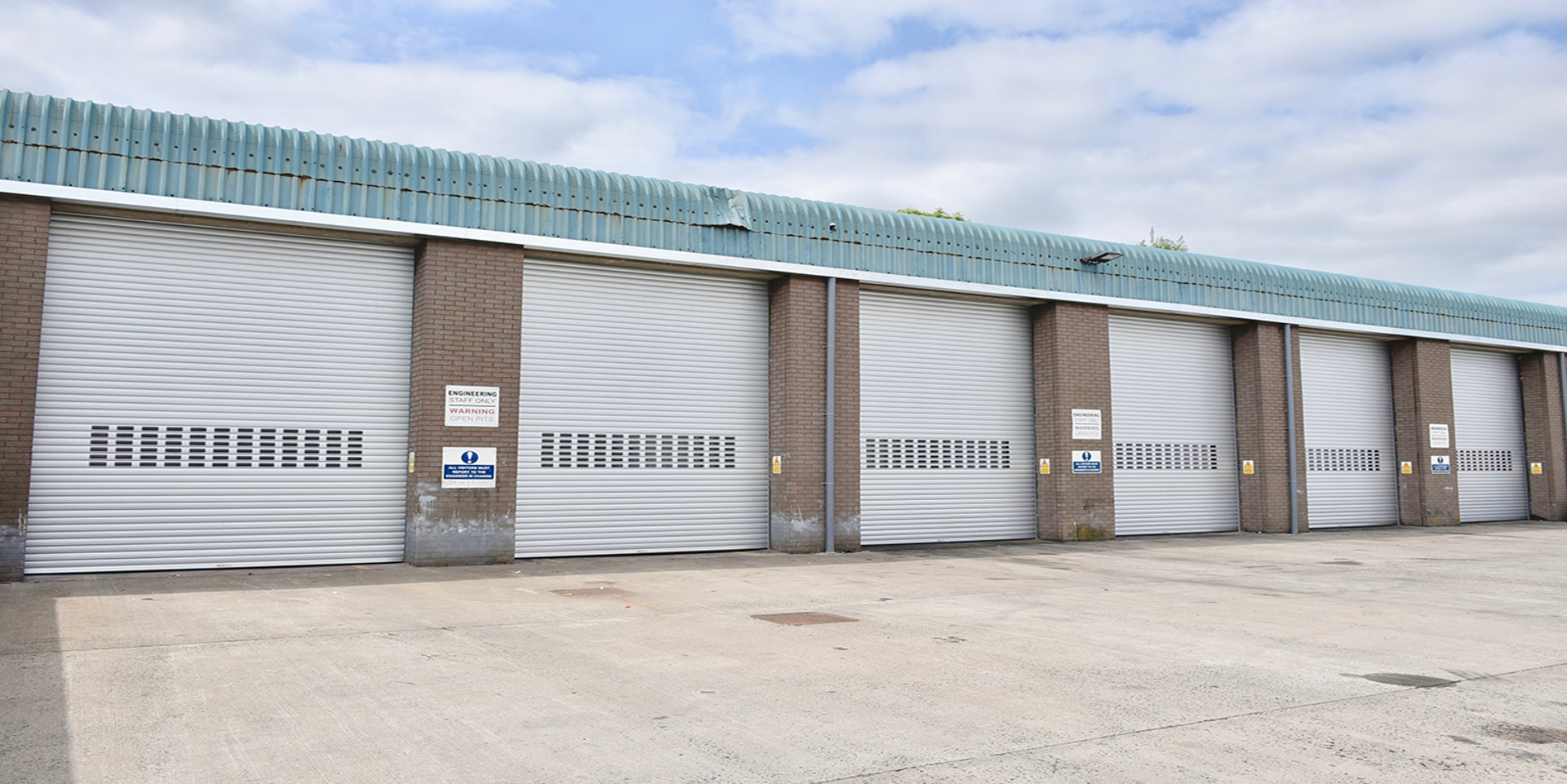 A row of closed industrial roller shutter doors on a brick building, with safety notices posted, under a partly cloudy sky.